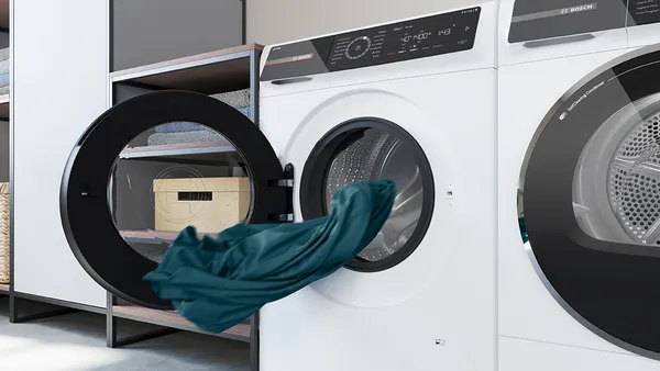 Una moderna lavatrice Bosch con porta trasparente che mostra un tamburo riempito d'acqua, accanto a un'asciugatrice e asciugamani ordinatamente impilati su uno scaffale.